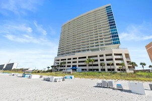 Hotel com vários andares e garagem, próximo a uma praia com espreguiçadeiras e guarda-sóis.