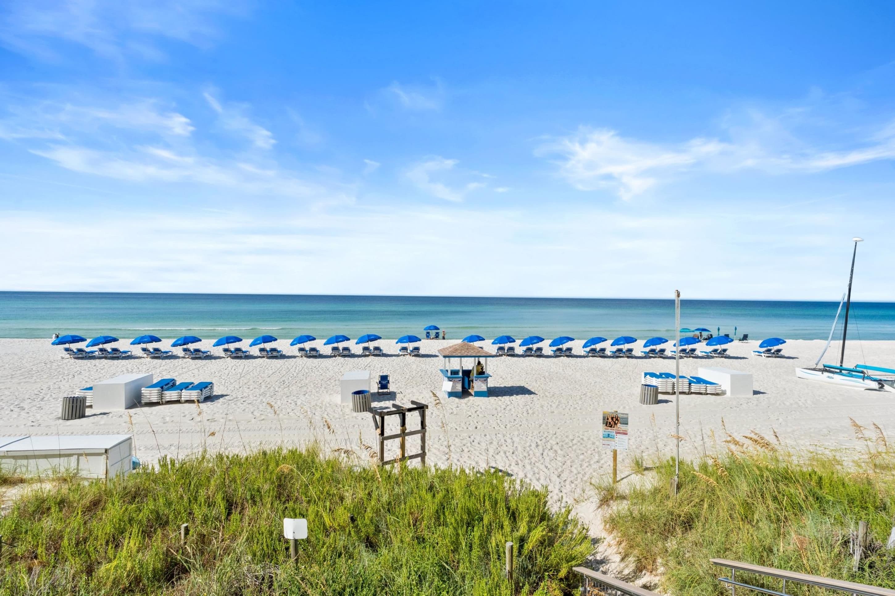 Una spiaggia con file di ombrelloni blu e lettini, un piccolo padiglione e una torretta per i bagnini.