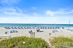 A beach with rows of blue umbrellas and lounge chairs, a small gazebo, and a lifeguard tower.