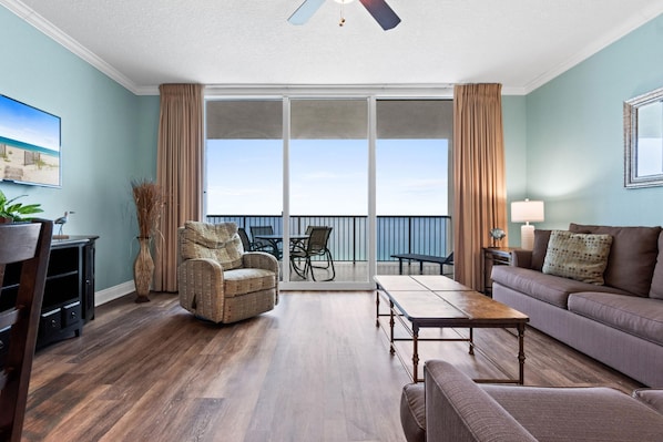 A living room with a sofa, armchair, coffee table, and a TV. There is a balcony with a view of the ocean.