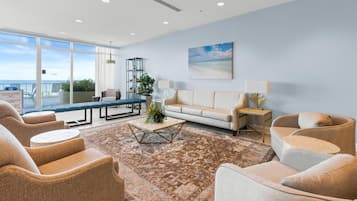 Ampla sala de estar com uma grande janela com vista para a praia, um sofá, poltronas, uma mesa de centro e uma mesa lateral com abajur.