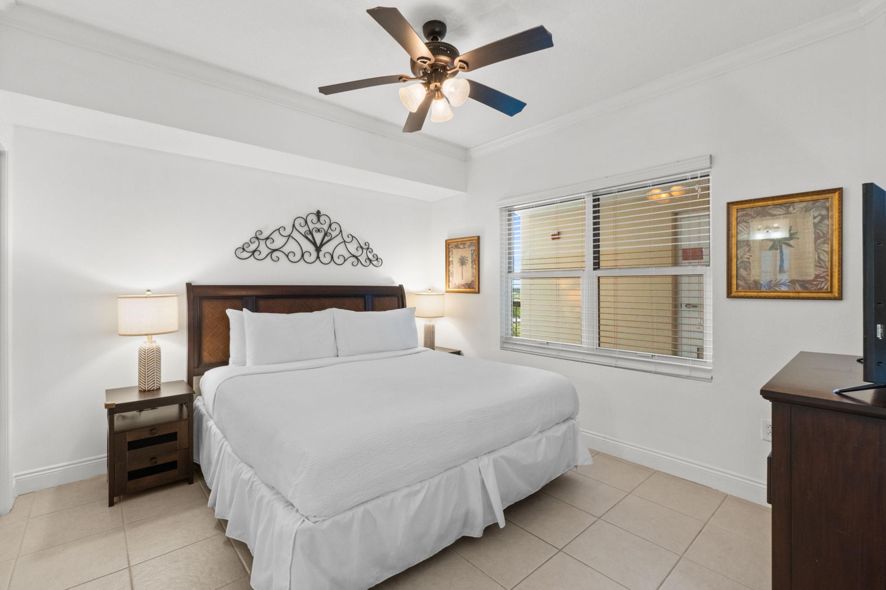 Une chambre à coucher comprenant un lit, des tables de chevet, un ventilateur de plafond, un élément décoratif mural, une fenêtre avec des stores et un téléviseur posé sur un meuble.