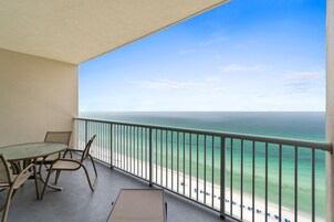 En balkon med bord og stole, med udsigt over en strand med klart vand og en fjerntliggende kystlinje.