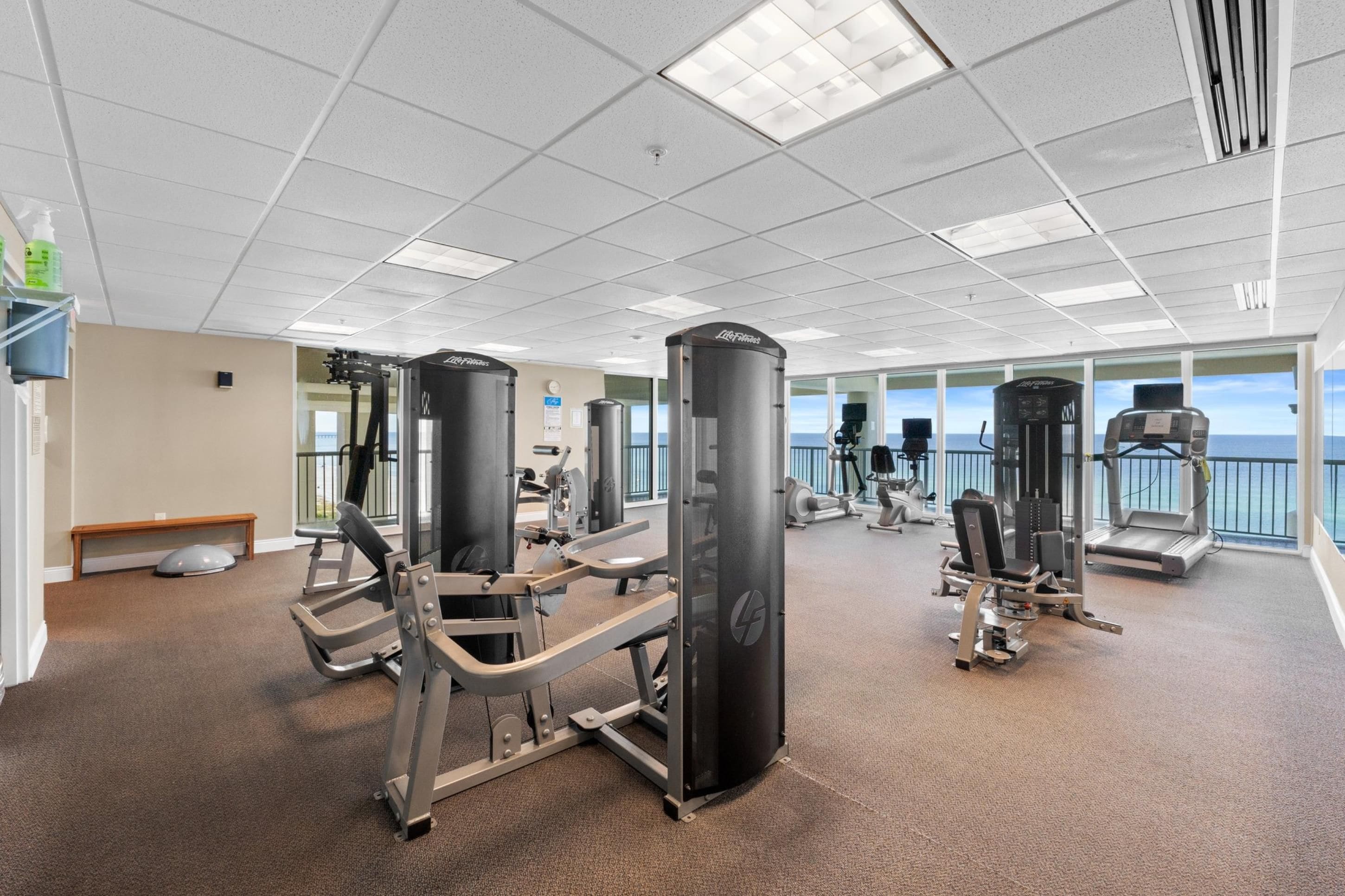 A gym with various exercise machines, a bench, and large windows offering a view of the ocean.
