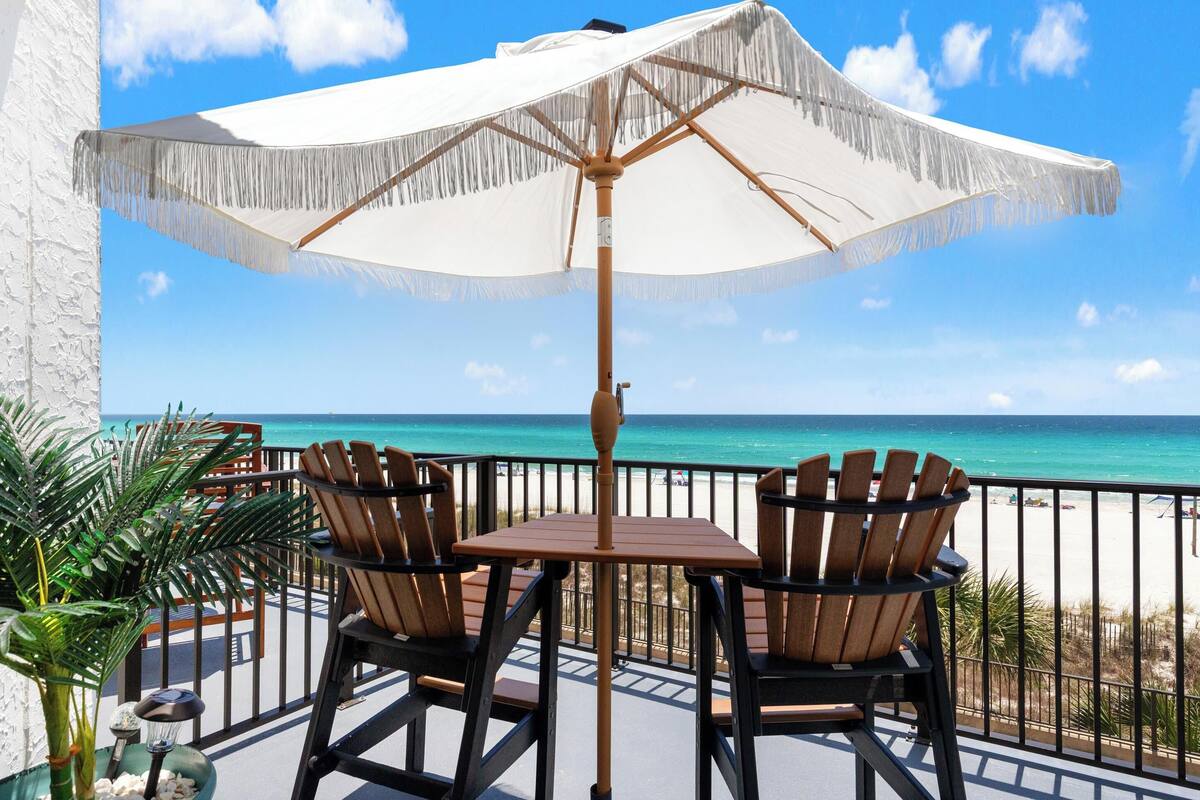 A balcony with a table and chairs, a white umbrella, and a view of the ocean.
