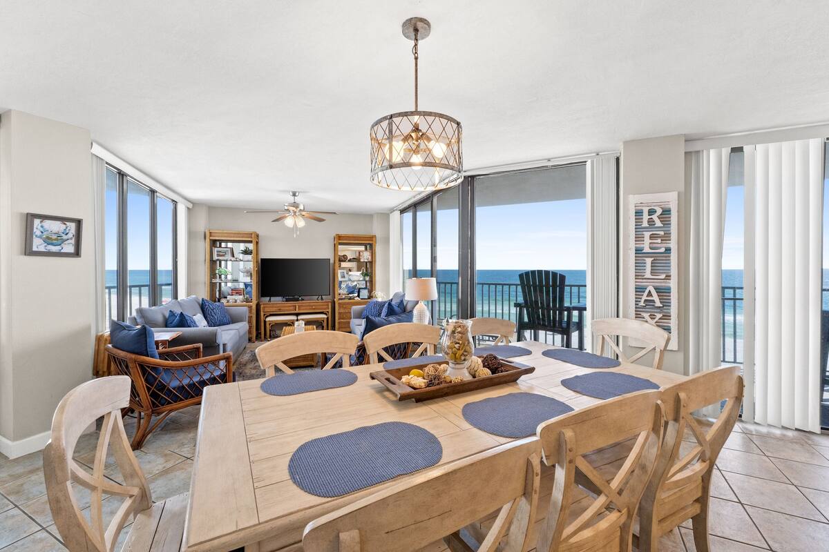 A spacious dining area with a wooden table and chairs, a chandelier, and a view of the ocean.