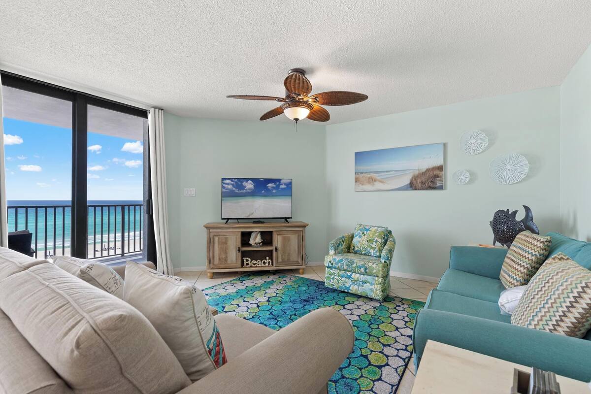 A living room with a view of the ocean, a ceiling fan, a television, a wooden cabinet, and a patterned rug.