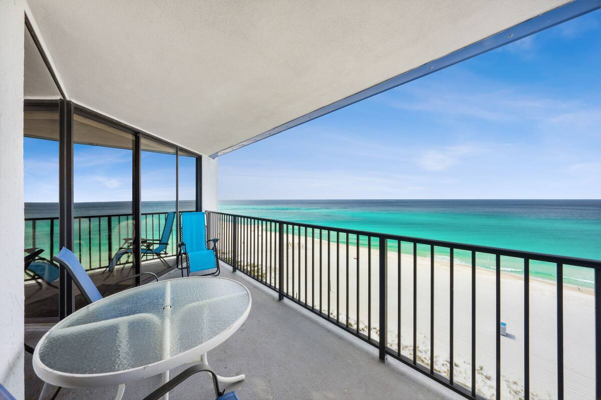 A balcony with a glass table and chairs overlooking a beach with clear turquoise water and a sandy shore.
