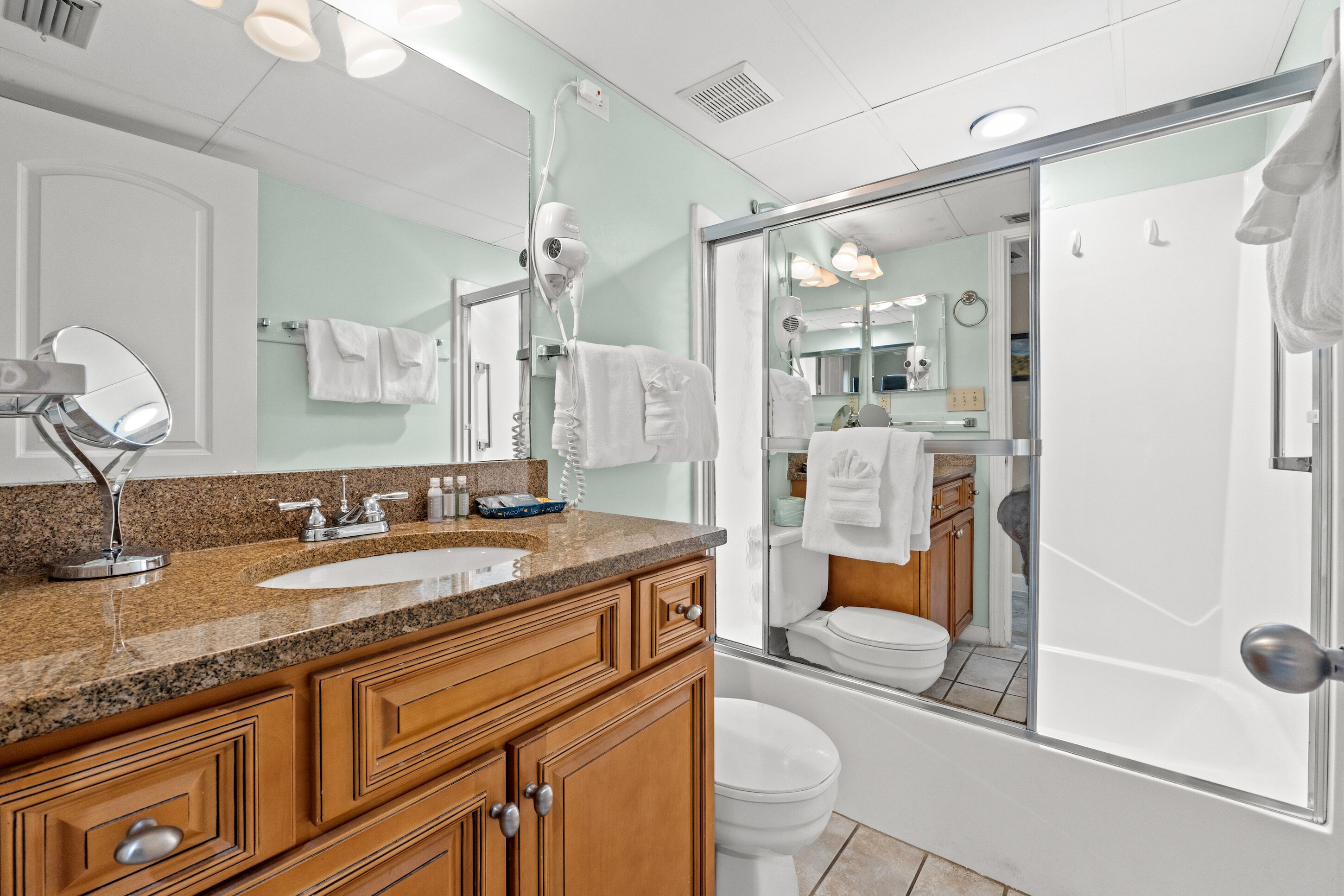 Een moderne badkamer met een granieten aanrechtblad, een grote spiegel, een haardroger en een douchecabine met glazen wanden.