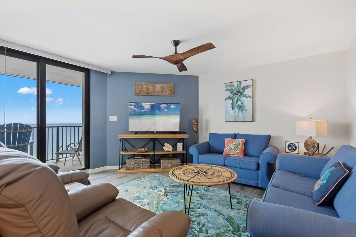 A living room with a blue sofa, a brown sofa, a ceiling fan, a TV, and a view of the ocean.