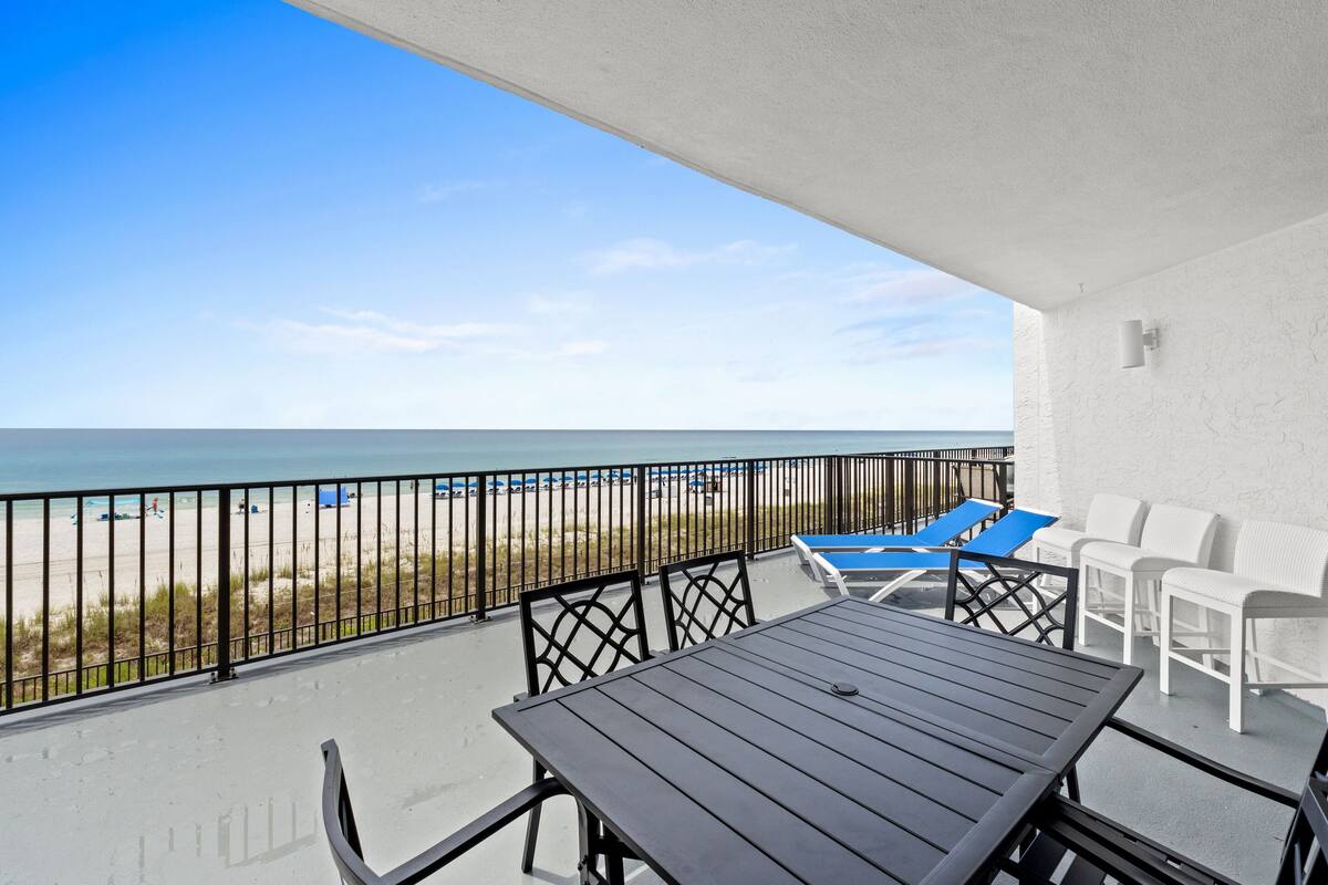 A balcony with a view of the beach, a table, and white chairs.