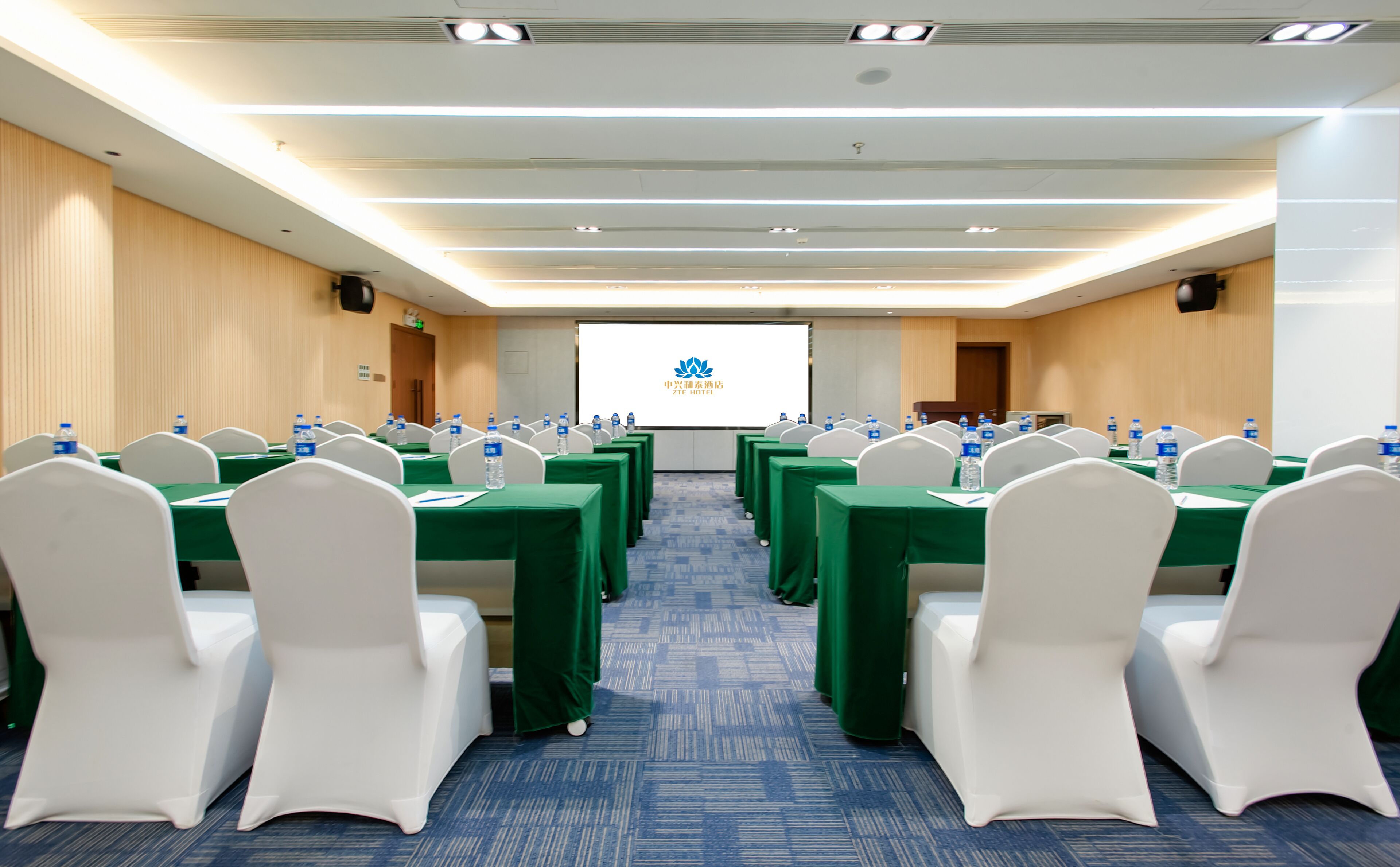 A conference room with rows of tables and chairs, water bottles on each table, and a presentation screen at the front.