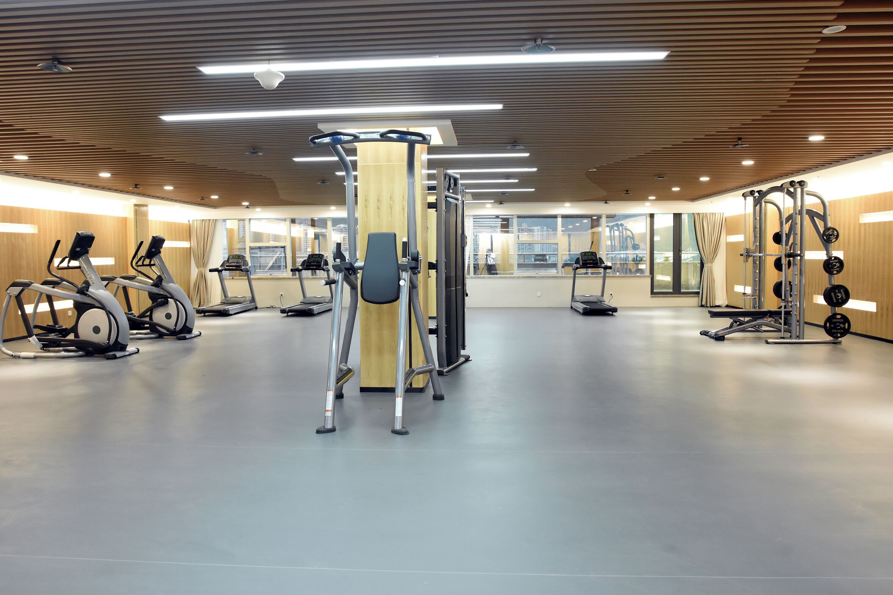 A modern gym with various exercise equipment, large windows, and a wooden ceiling.
