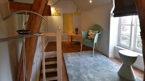 Un escalier avec des poutres en bois, une petite table avec une lampe, une chaise avec un coussin orné de motifs de papillons et une fenêtre donnant sur un paysage verdoyant.