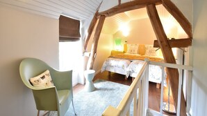 Une chambre avec un plafond en pente, un lit recouvert d’une literie à motifs floraux, un fauteuil vert et une petite table avec une lampe.