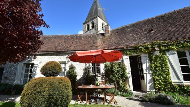 Un bâtiment en pierre avec une tour, un jardin avec une table et des chaises, et un parasol rouge.