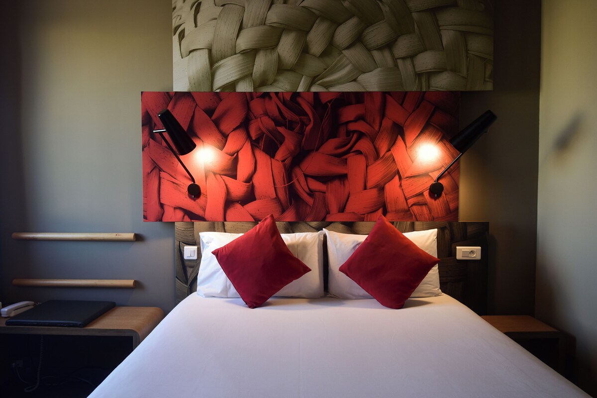 Una habitación de hotel con una cama, almohadas rojas, cabecera con diseño tejido, mesitas de noche y una lámpara de pared.