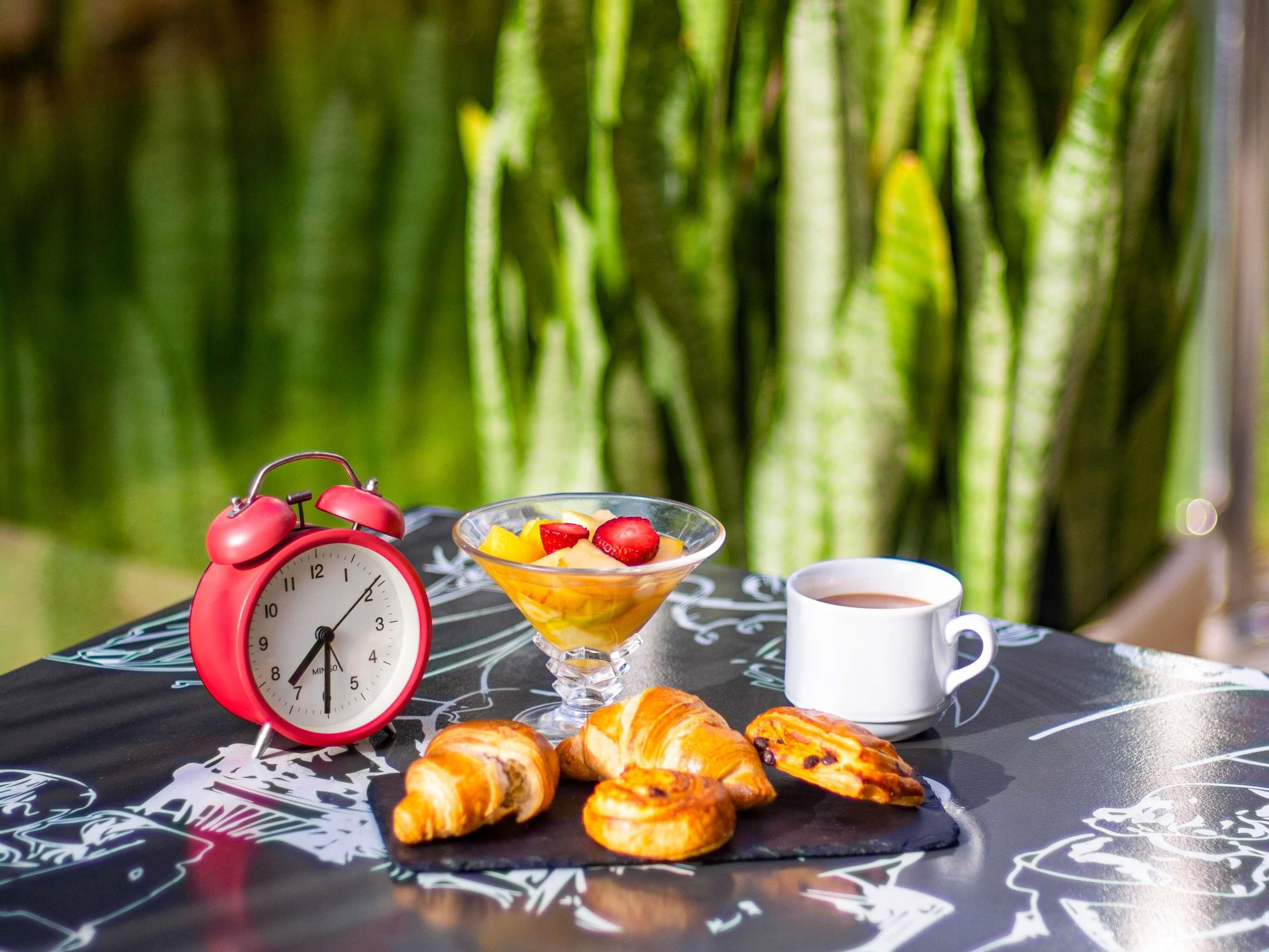A table with a red alarm clock, a bowl of fruit, a cup of coffee, and croissants.
