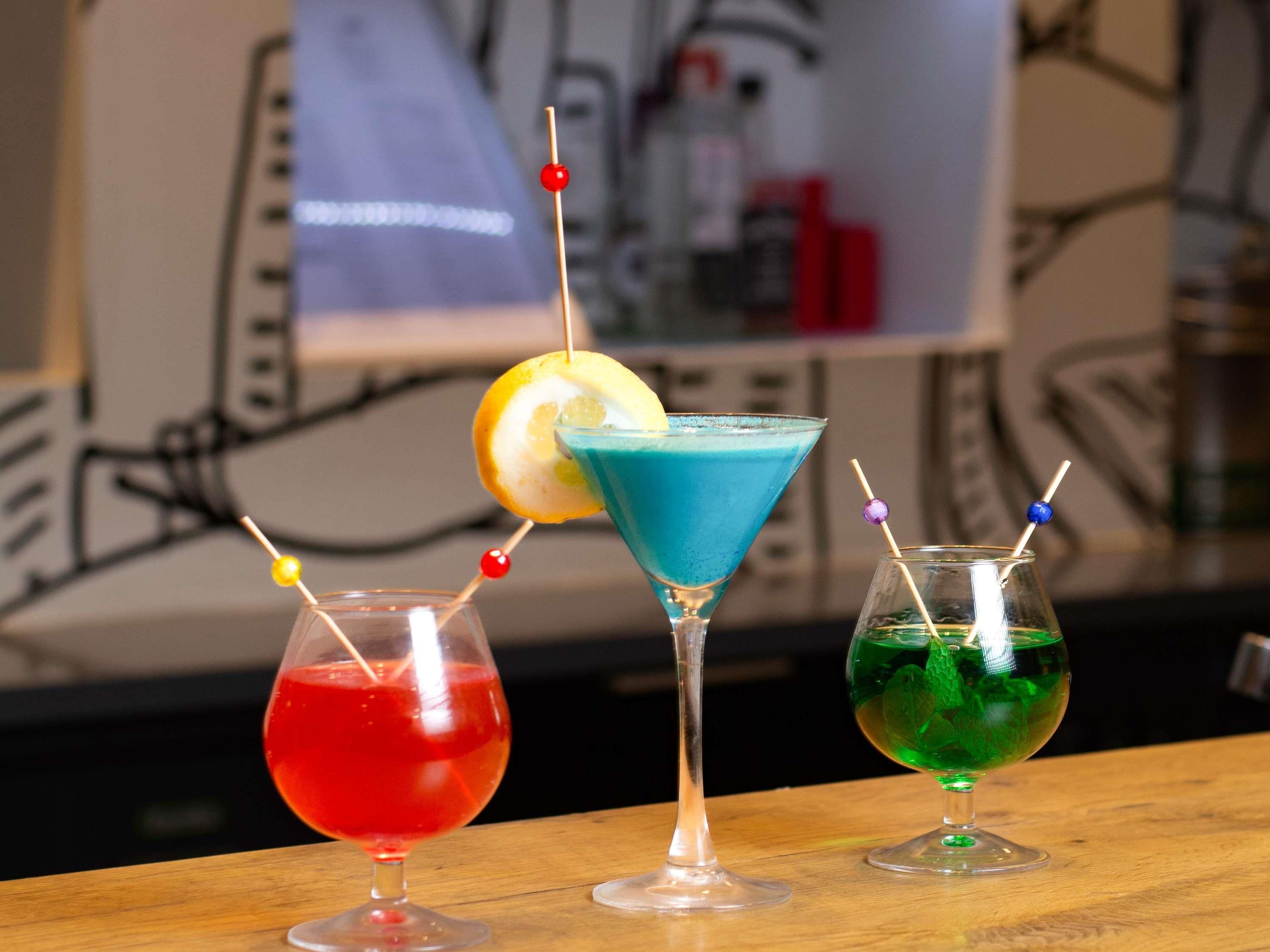 Trois cocktails servis dans des verres à martini, décorés de garnitures colorées, sur le comptoir en bois d’un bar.