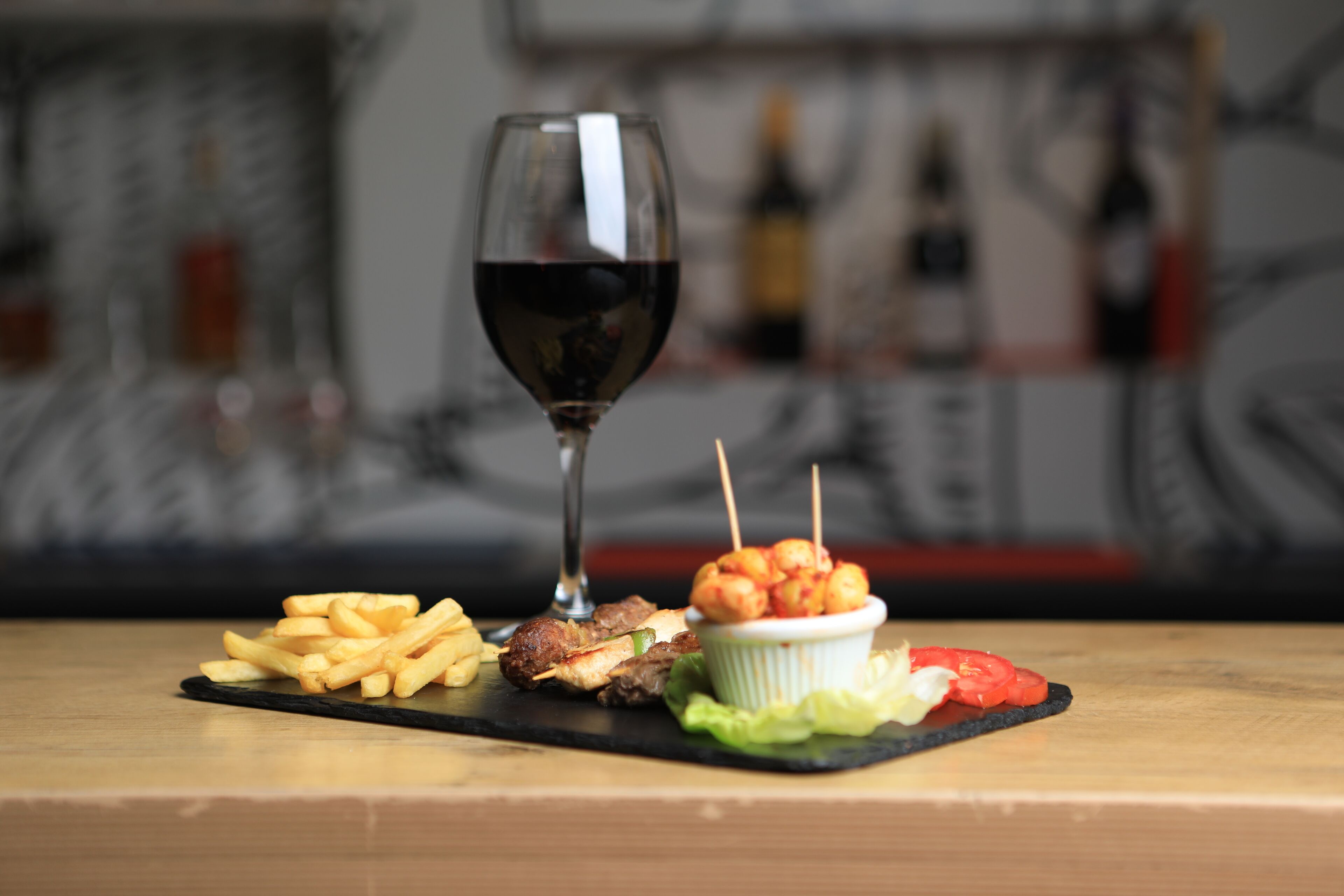 A meal with fries, meat skewers, and a salad on a black slate plate, accompanied by a glass of red wine.