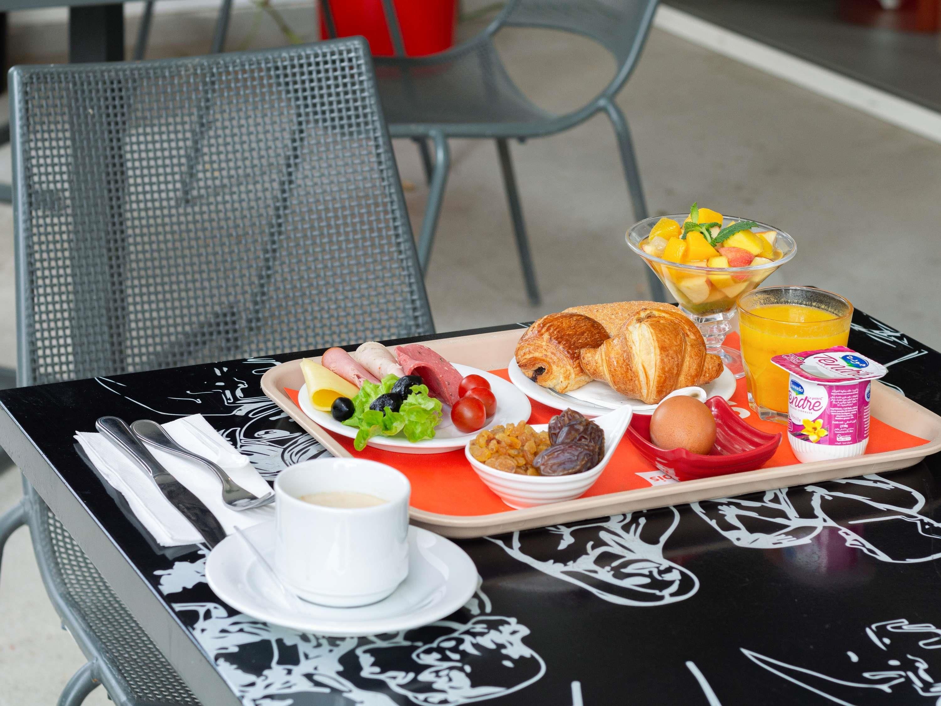 Bandeja de desayuno con cruasanes, frutas, yogur y una taza de café sobre una mesa con estampado blanco y negro.