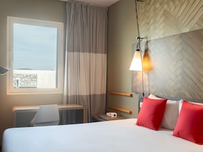 A hotel room with a bed, a desk, a chair, a window with curtains, a wall-mounted lamp, and a herringbone-patterned wall. - ibis Rabat Agdal (Rabat)