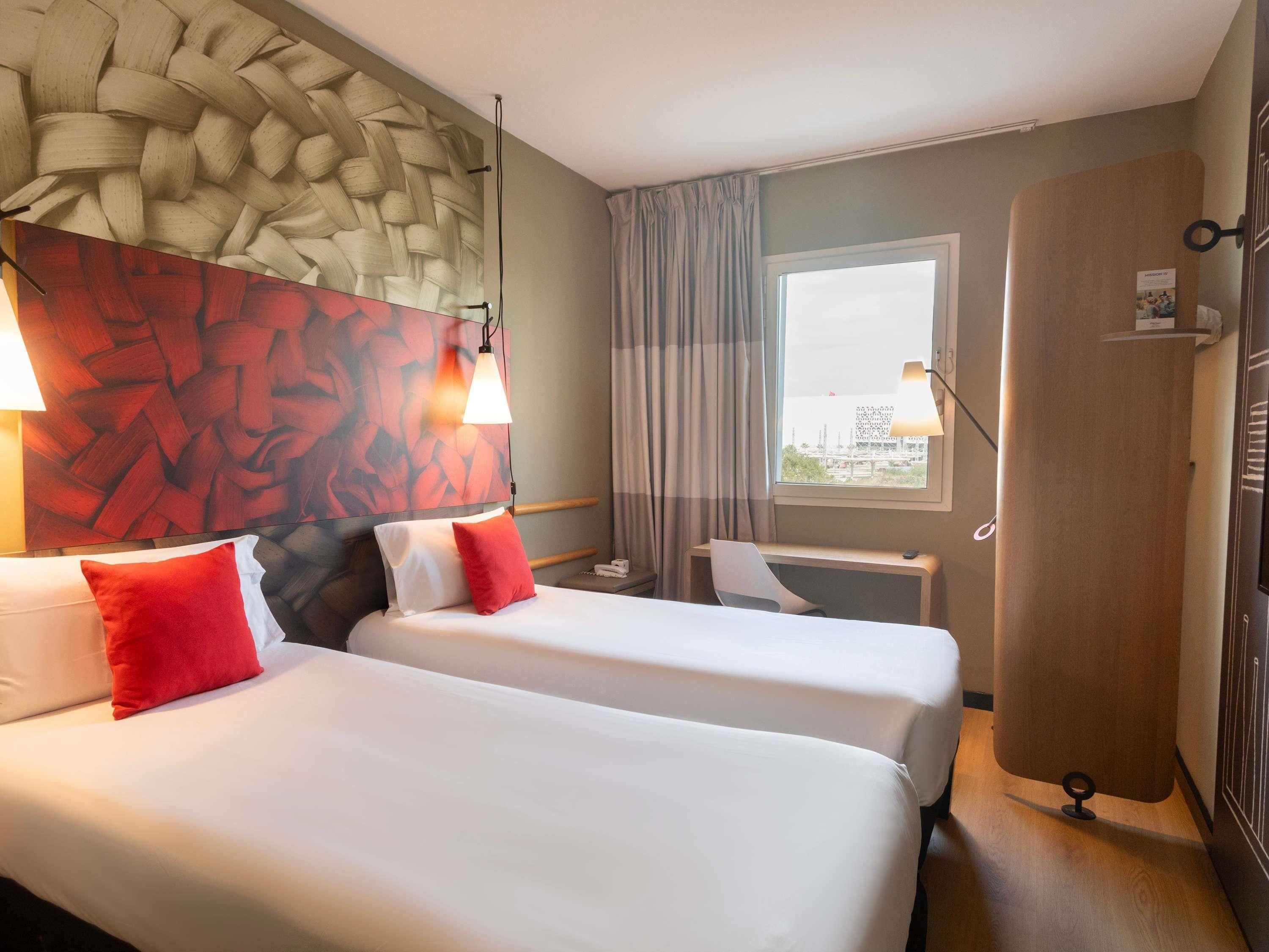 A hotel room with two beds, a large window, a desk, and a wall with a woven texture.