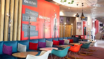 A modern interior with a red and grey wall featuring 'ibis kitchen' signage, a seating area with colorful sofas and tables, and a ceiling with hanging plants.