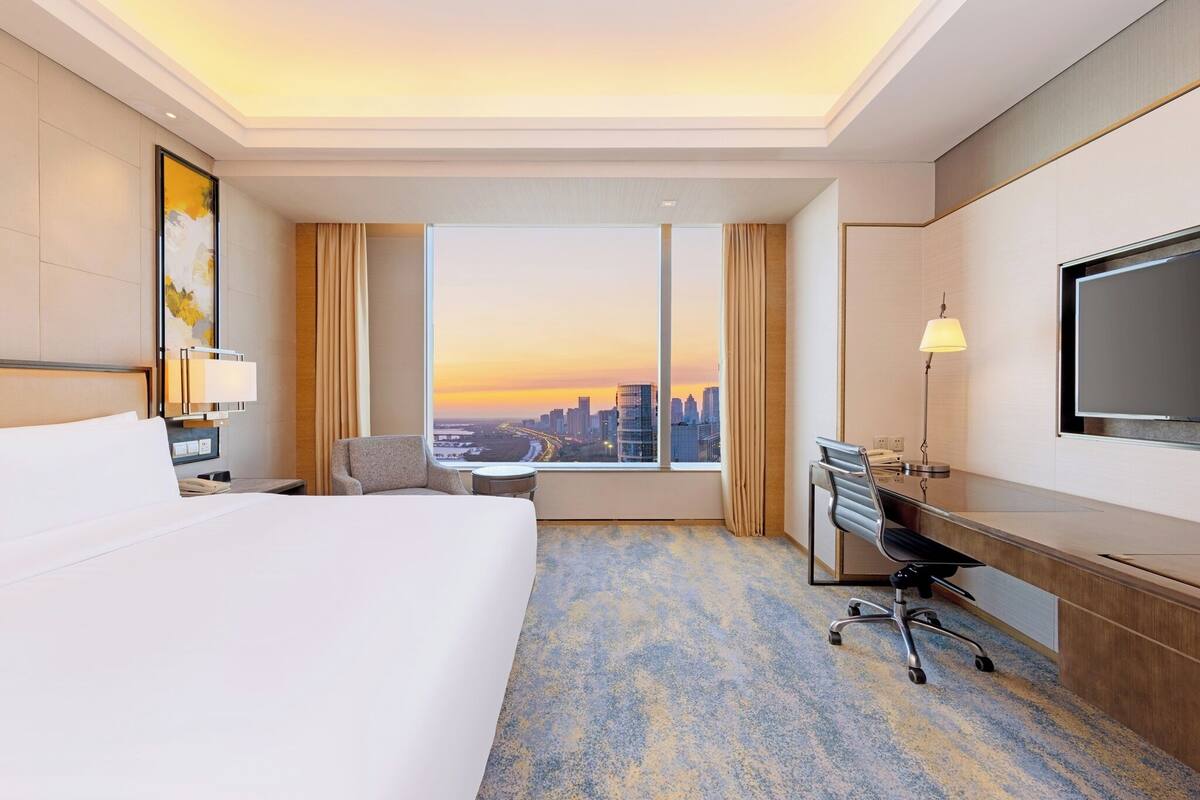 A hotel room with a large bed, a desk, a chair, a TV, and a window with a city view.