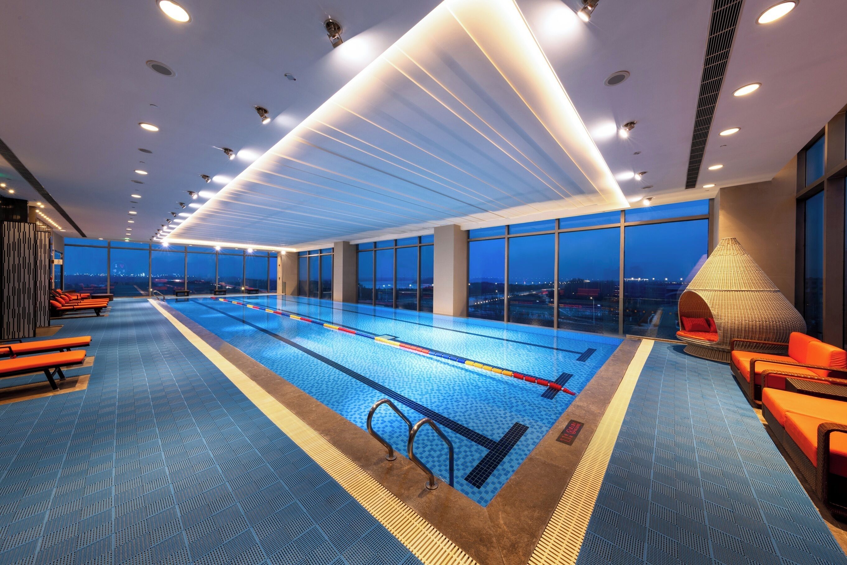 Piscina interior moderna com marcações nítidas nas pistas, piso de azulejos azuis e vista da cidade ao entardecer.