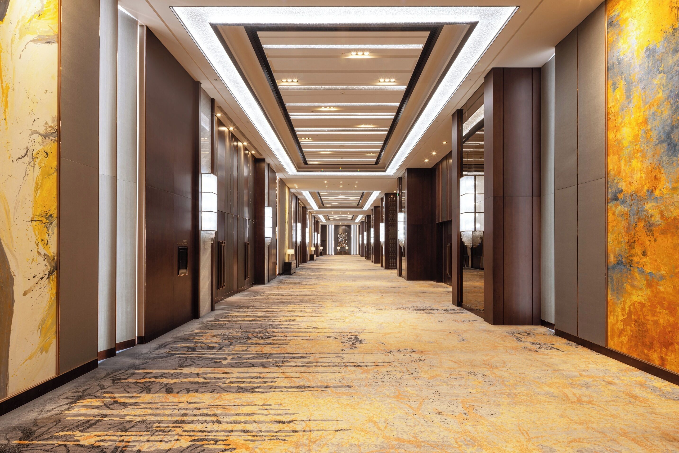 Un pasillo largo y estrecho con alfombra estampada, iluminación empotrada y paneles de madera en las paredes.