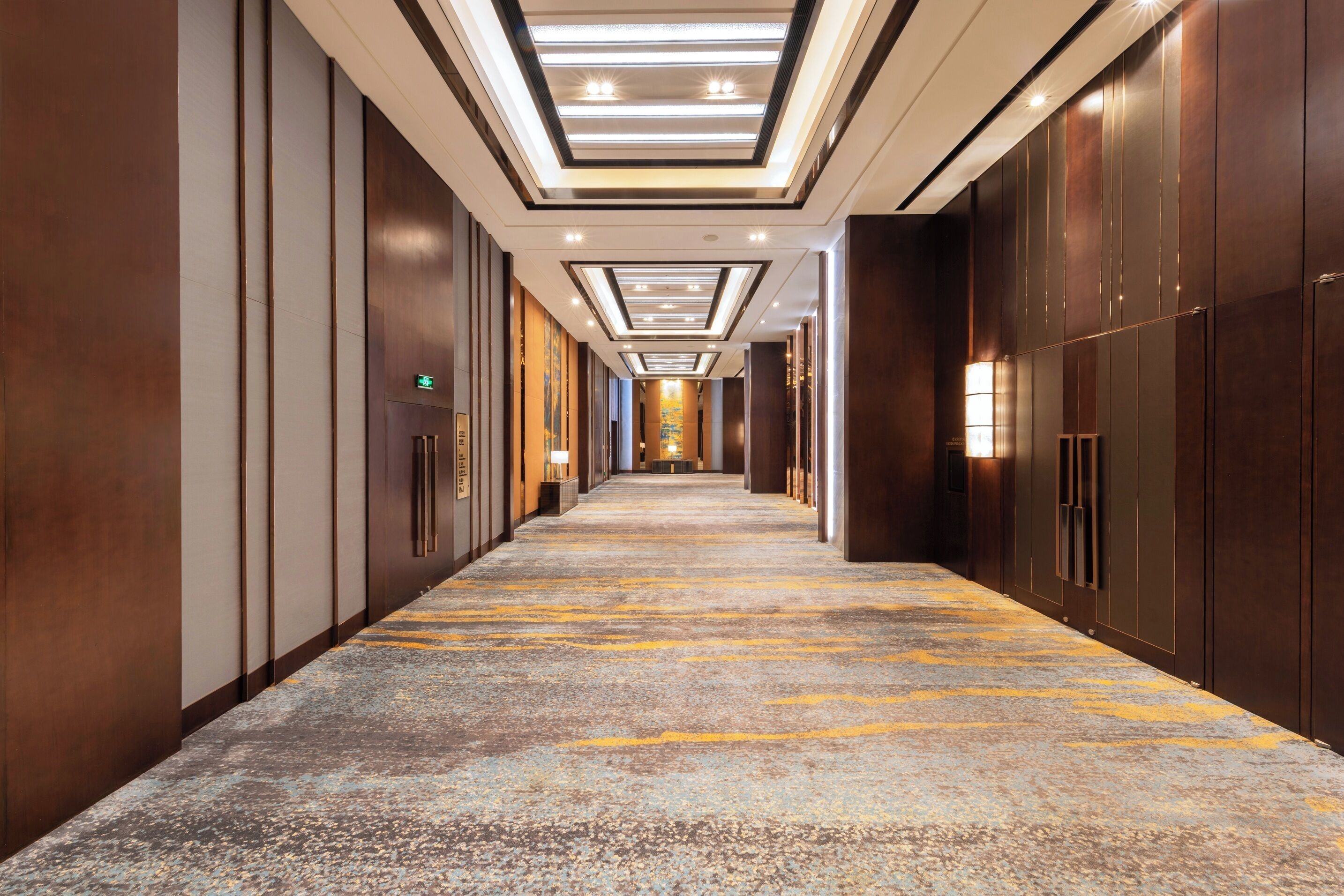 A long, narrow hotel corridor with a patterned carpet, wooden walls, and recessed lighting.
