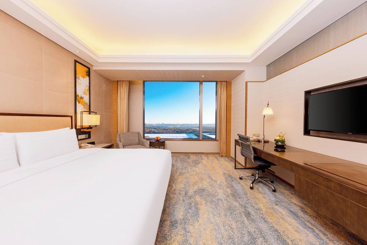 A hotel room with a large bed, a desk, a chair, a TV, and a view of the cityscape through the window.