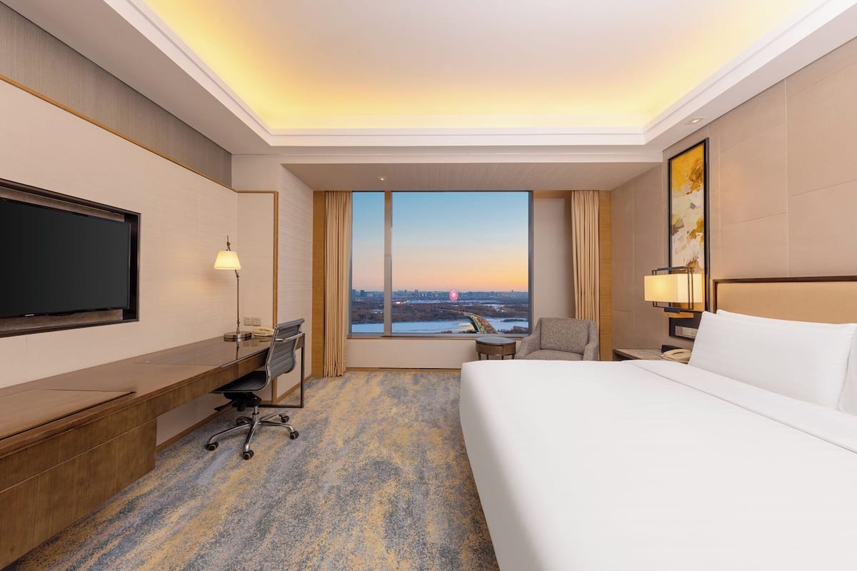 A hotel room with a large bed, a desk with a chair, a television, and a view of a cityscape at sunset.