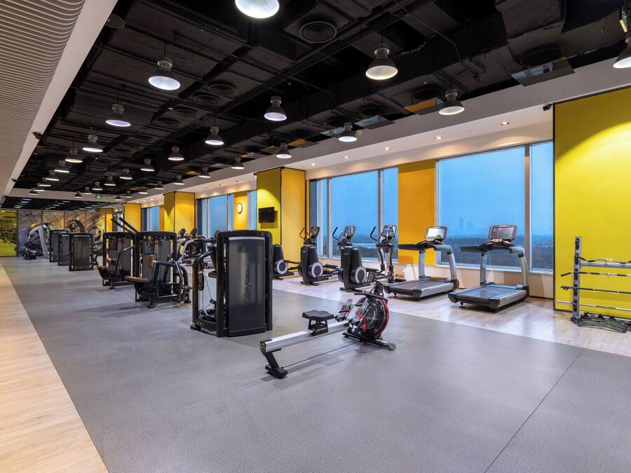 A modern gym with cardio machines, weight stations, and a large window offering a city view.