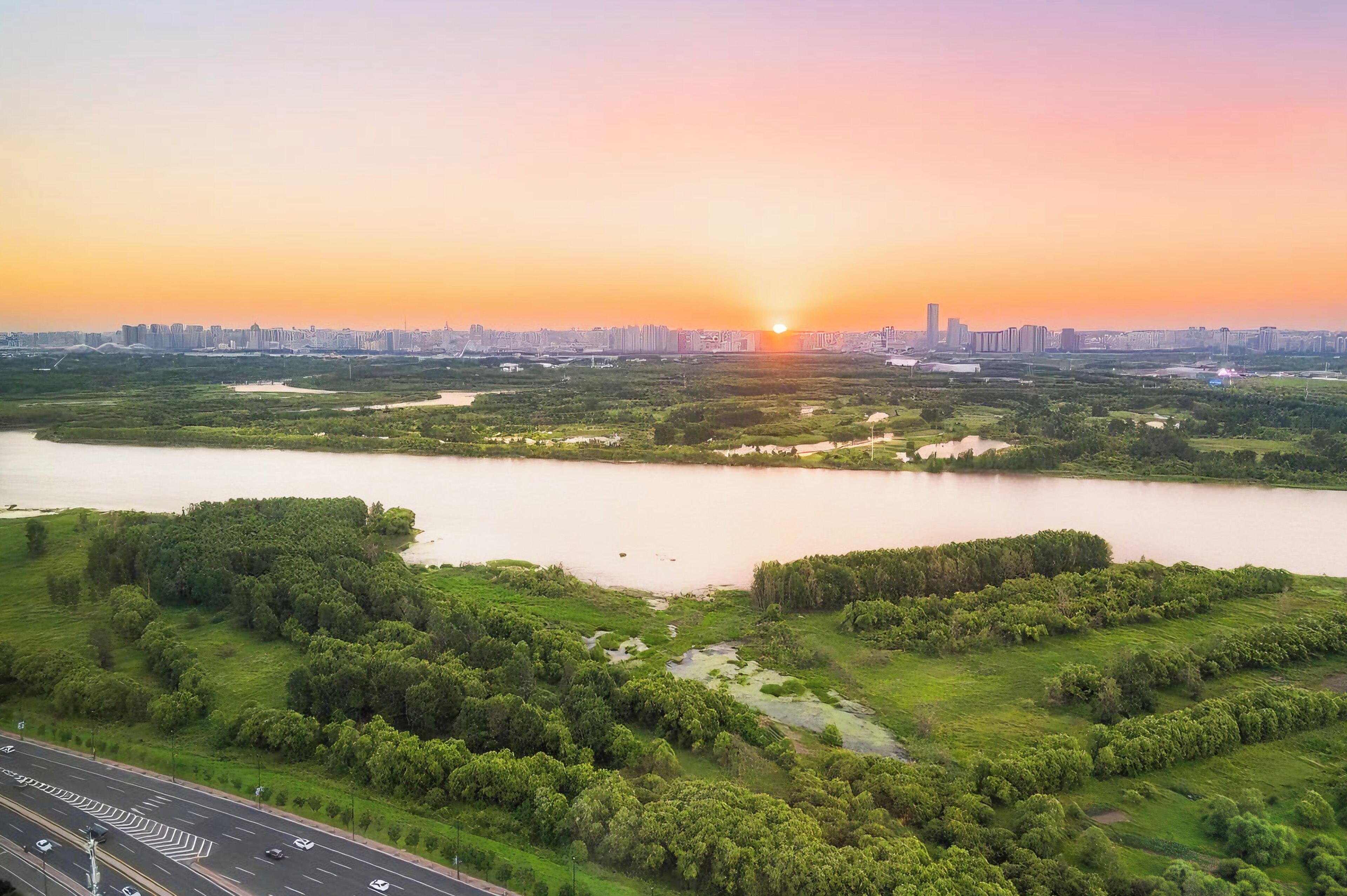 Paisagem urbana ao pôr do sol, com um rio, vegetação e uma rodovia.