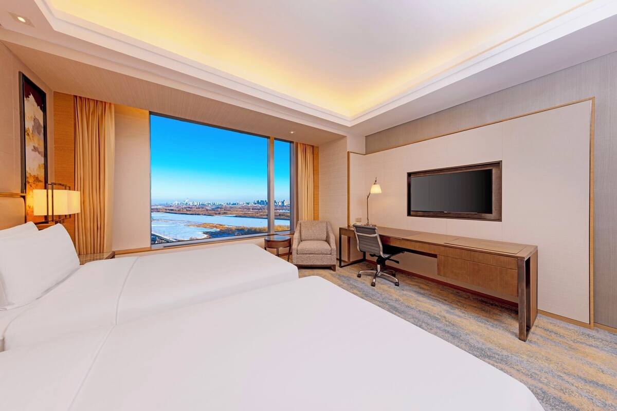 A hotel room with a large bed, a desk, a chair, a TV, and a view of a cityscape.
