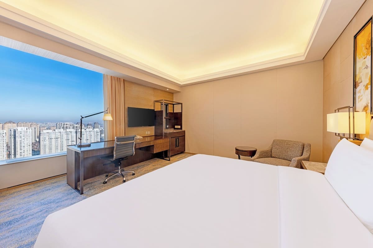 Quarto de hotel com uma cama grande, uma escrivaninha com cadeira, televisão e vista para a cidade.
