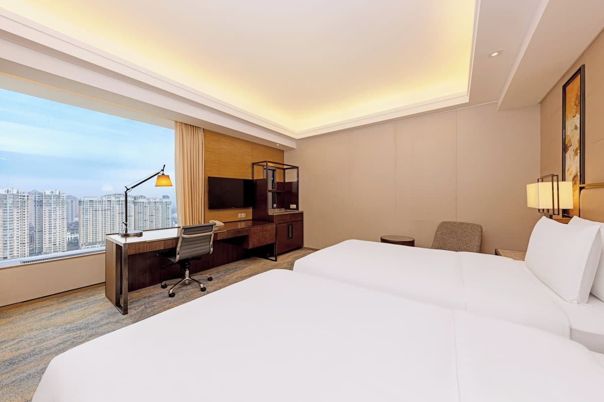Quarto de hotel com escrivaninha, duas camas, uma cadeira, abajur e uma janela grande com vista para a cidade.