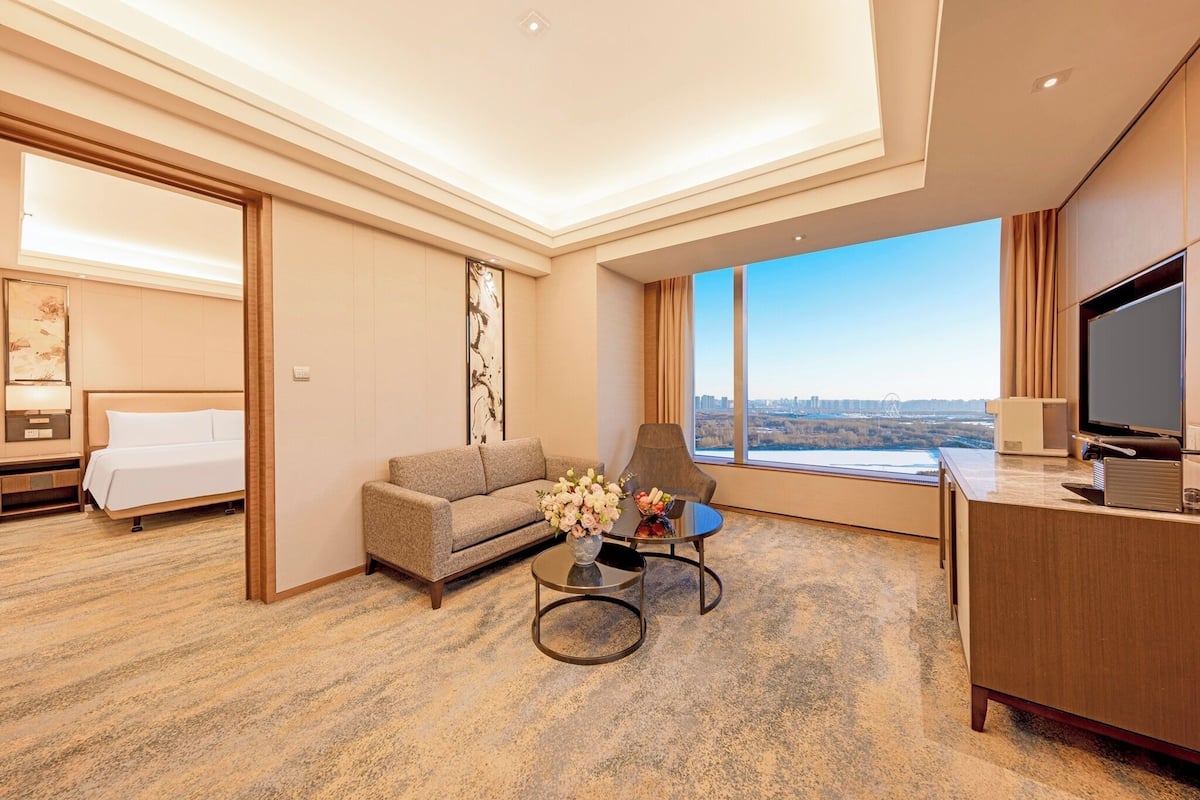 A hotel room with a bed, sofa, armchair, coffee table, TV, and a large window with a city view.