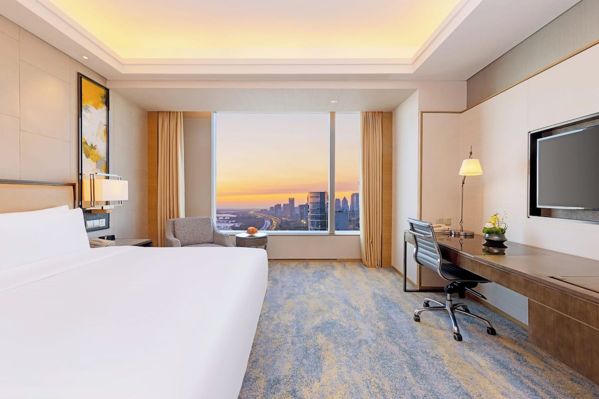 A hotel room with a large bed, a desk, a chair, a TV, and a view of the city skyline.