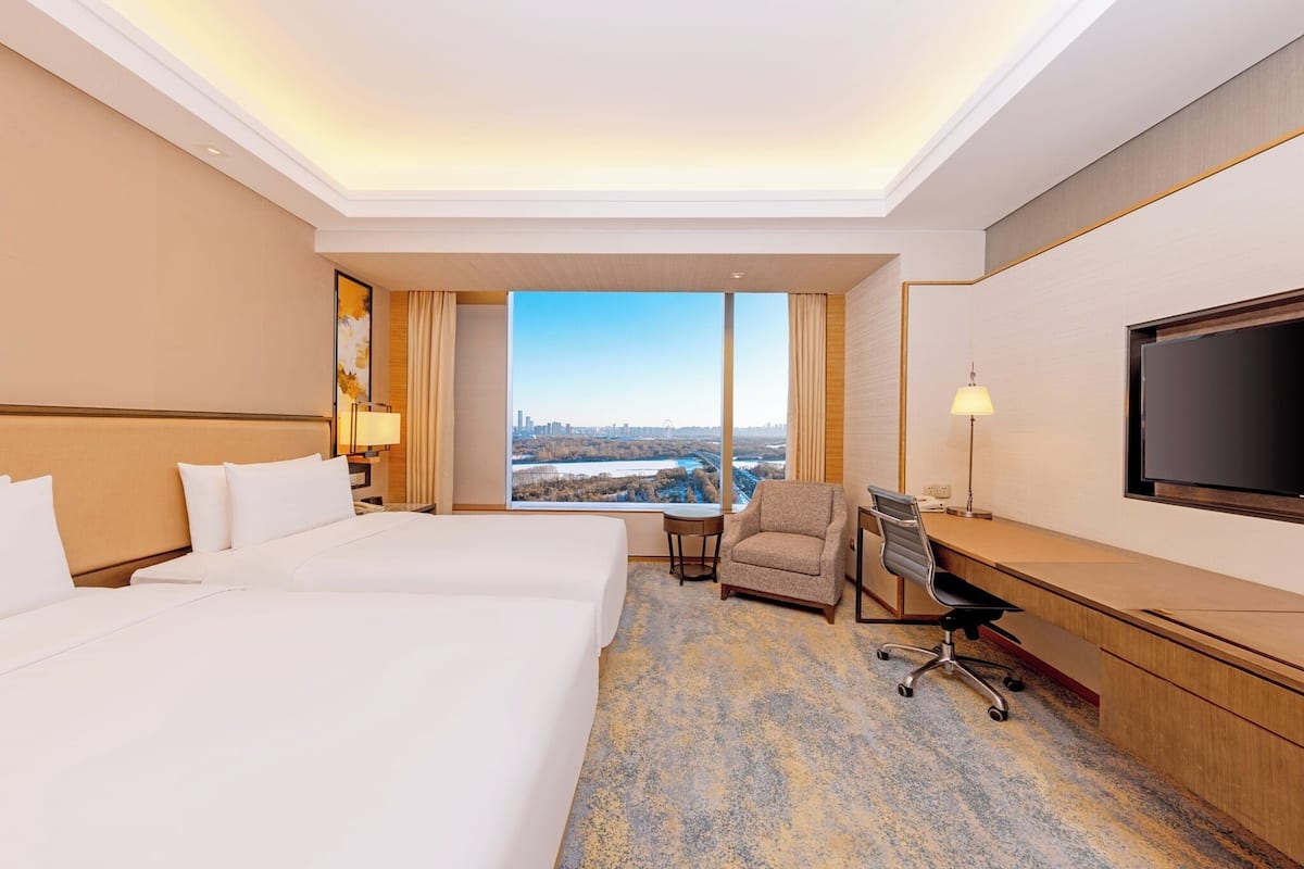 A hotel room with two beds, a desk, a chair, a TV, and a large window with a city view.