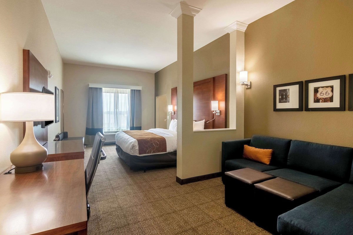 A hotel room with a bed, a sofa, a desk with a lamp, and framed pictures on the wall.