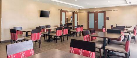 A hotel dining area with multiple tables and chairs, a flat-screen TV mounted on the wall, and a sliding door closet.