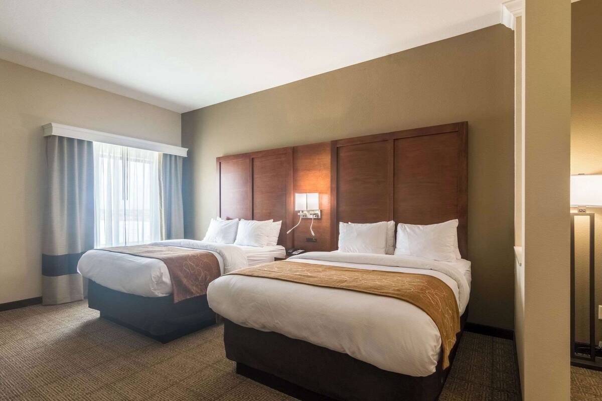 A hotel room with two beds, a large wooden headboard, a window with curtains, and a bedside lamp.