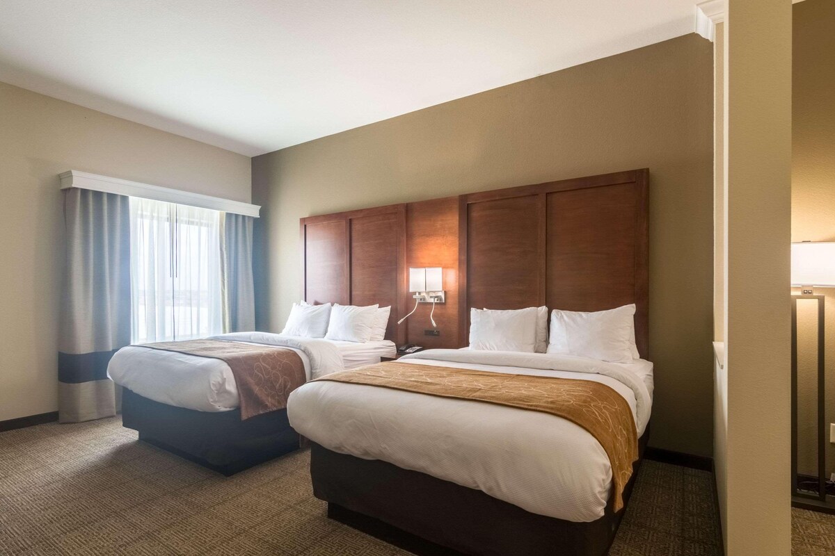 Habitación de hotel con dos camas, un ventanal con cortinas, un cabecero de madera y una lámpara de noche.