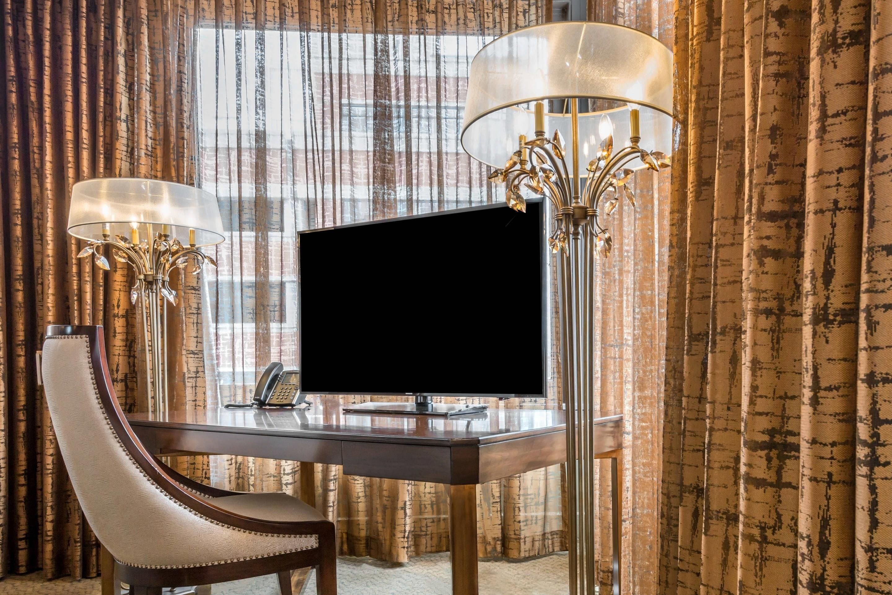A hotel room with a desk, a television, two lamps, and a chair.