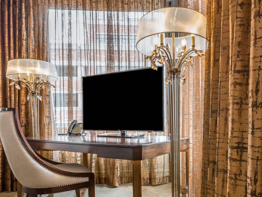 A hotel room with a desk, a television, two lamps, and a chair.