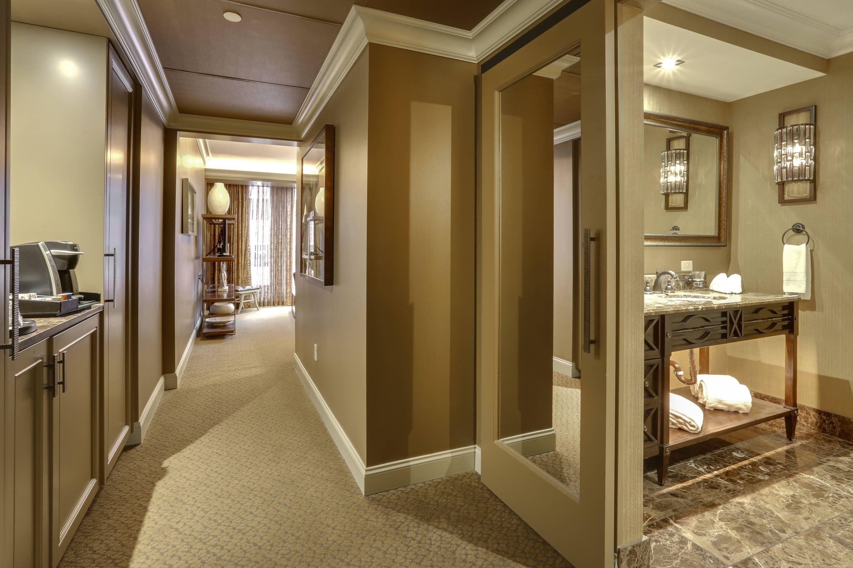 A hotel hallway with a carpeted floor, a wooden door, a vanity with a mirror, and a towel rack.