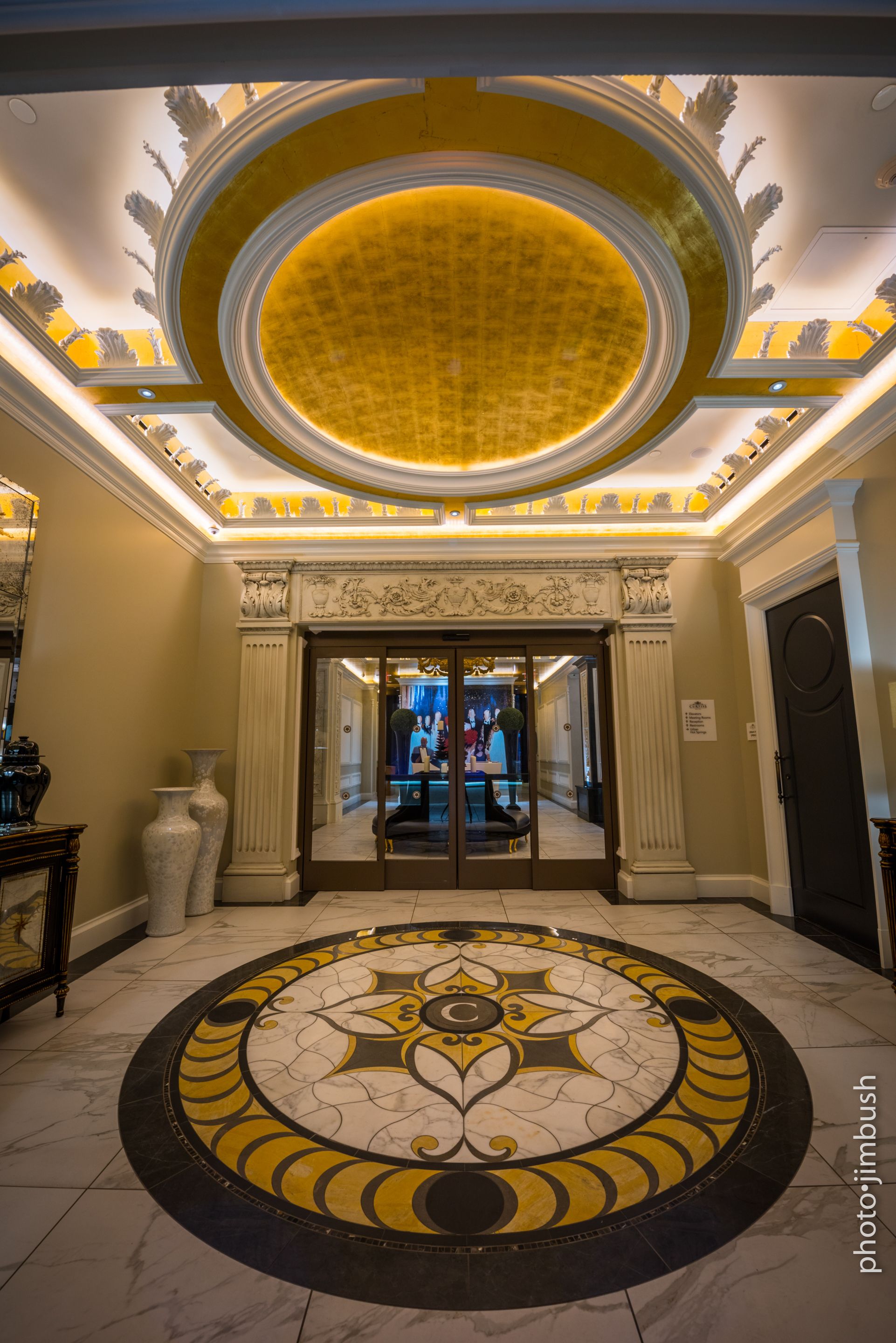 A grand lobby with a large circular mosaic floor, a high ceiling with ornate decorations, and a glass door entrance.